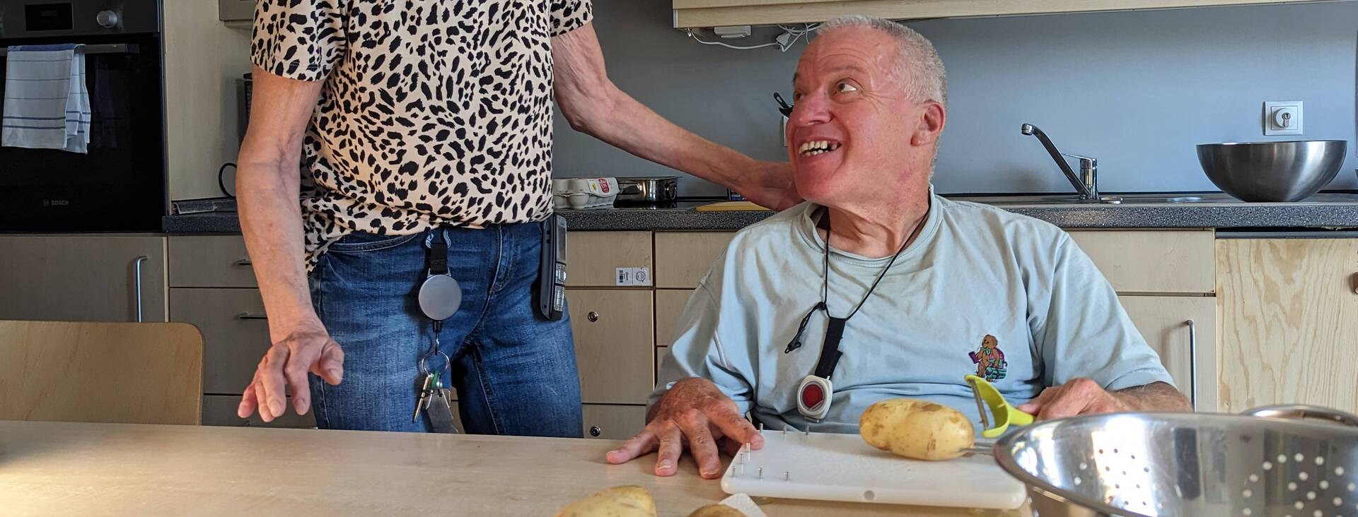 Eine Betreuerin hilft einem Wohnheim Bewohner bei dem Kartoffel schälen, beide lächeln fröhlich..